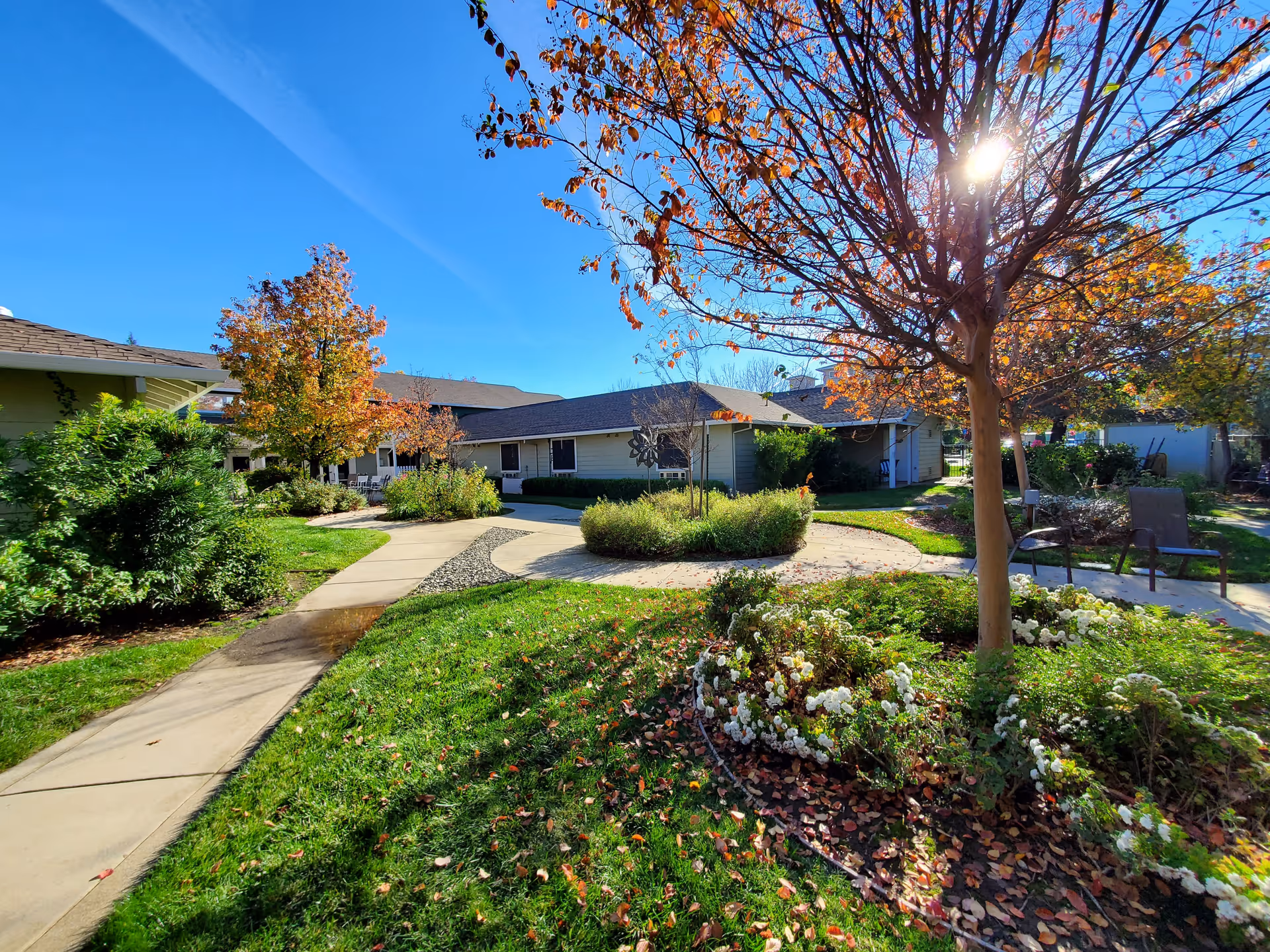 Outdoor garden area at Sundial Assisted Living with a paved walkway, green grass, flower beds, and trees with autumn leaves under a clear blue sky. There are chairs placed near the walkway and sunlight filtering through the branches of a tree.