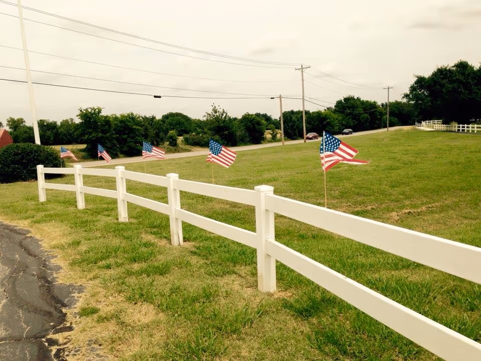 A grassy outdoor area with a white wooden fence running alongside a paved path. Several small American flags are placed in the grass near the fence. Trees and utility poles are visible in the background under a cloudy sky.