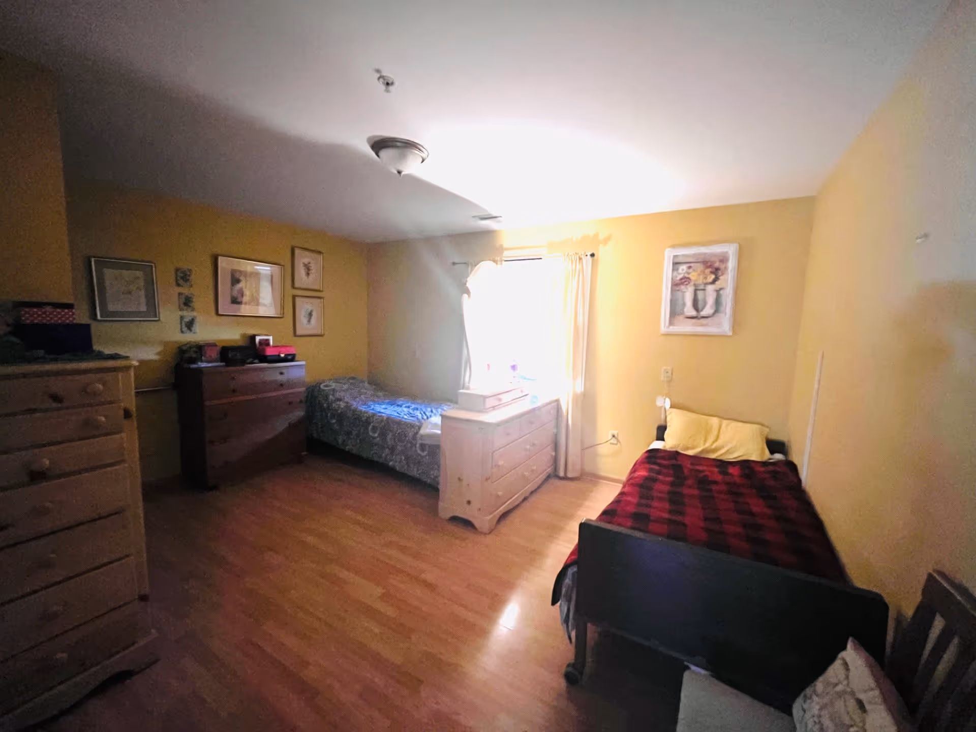A cozy bedroom featuring two beds, one with a red and black checkered blanket, and a dresser. The walls are painted yellow, and there are framed artworks and a window with curtains allowing natural light.