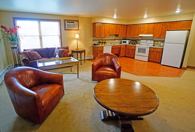 A cozy living area combined with a kitchen. The living area has two brown leather armchairs, a round wooden coffee table, a sofa with cushions, a glass coffee table, a floor lamp, and a window with blinds. The kitchen features wooden cabinets, a white refrigerator, stove, microwave, and dishwasher, with a wood floor section distinct from the carpeted living area.