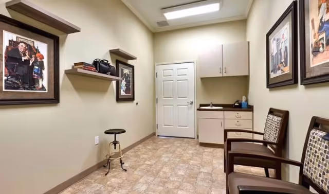 A small, clean room with beige walls and tiled floor, featuring a white door at the back, a cabinet with a countertop and sink on the right side, two wooden chairs with patterned cushions, a black adjustable stool, two floating shelves with books and a camera, and framed pictures on the walls.