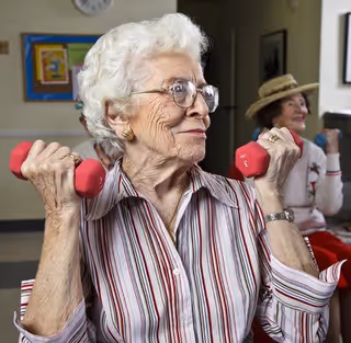 Two elderly women exercising indoors, each holding a pair of small red dumbbells. The woman in the foreground has white hair, glasses, and is wearing a striped shirt. The woman in the background is wearing a hat and a white sweater with red accents.