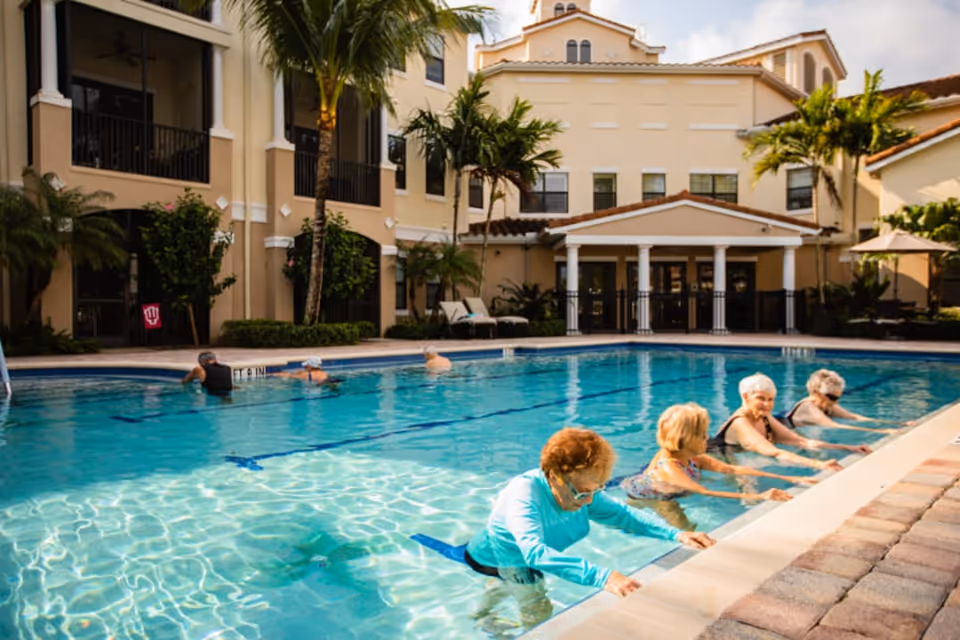 Several seniors exercising in an outdoor swimming pool in front of a multi-story senior living building with palm trees.
