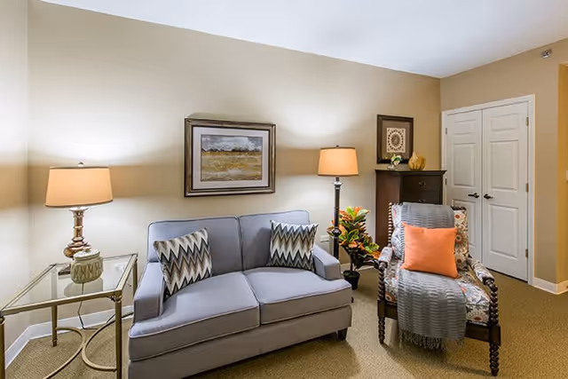 A cozy living room area with a light gray loveseat adorned with two patterned pillows, a wooden armchair with a floral cushion, an orange throw pillow, and a gray blanket. There are two table lamps on either side, one on a glass side table and the other on a small wooden cabinet. The walls are beige with framed artwork hanging above the loveseat and cabinet. A double white door is visible in the background.
