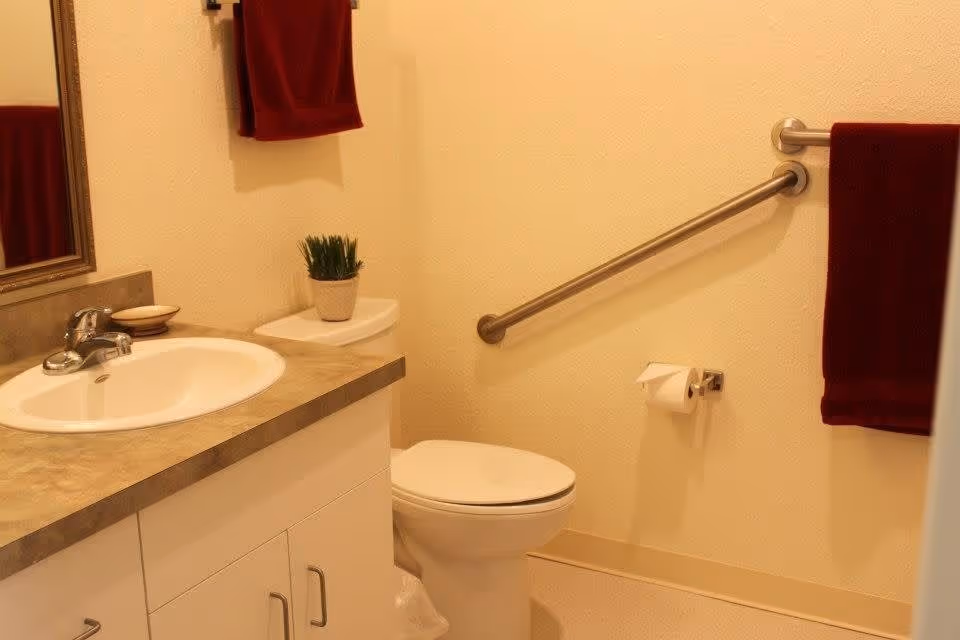 A clean bathroom with a white sink and countertop, a toilet with a small potted plant on the tank, a grab bar on the wall next to the toilet, and two red towels hanging on the walls.