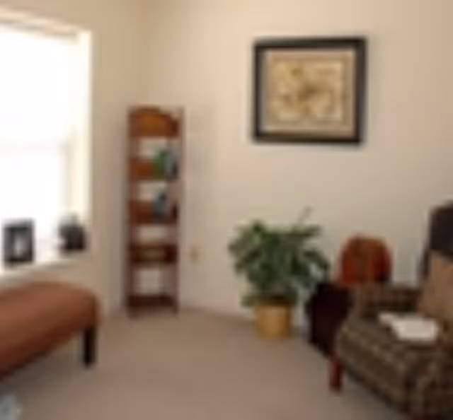 A cozy living room area with a patterned armchair, a small wooden side table with a decorative item, a potted plant on the floor, a tall wooden bookshelf with books and decor, a cushioned bench near a window, and a framed artwork hanging on the wall.