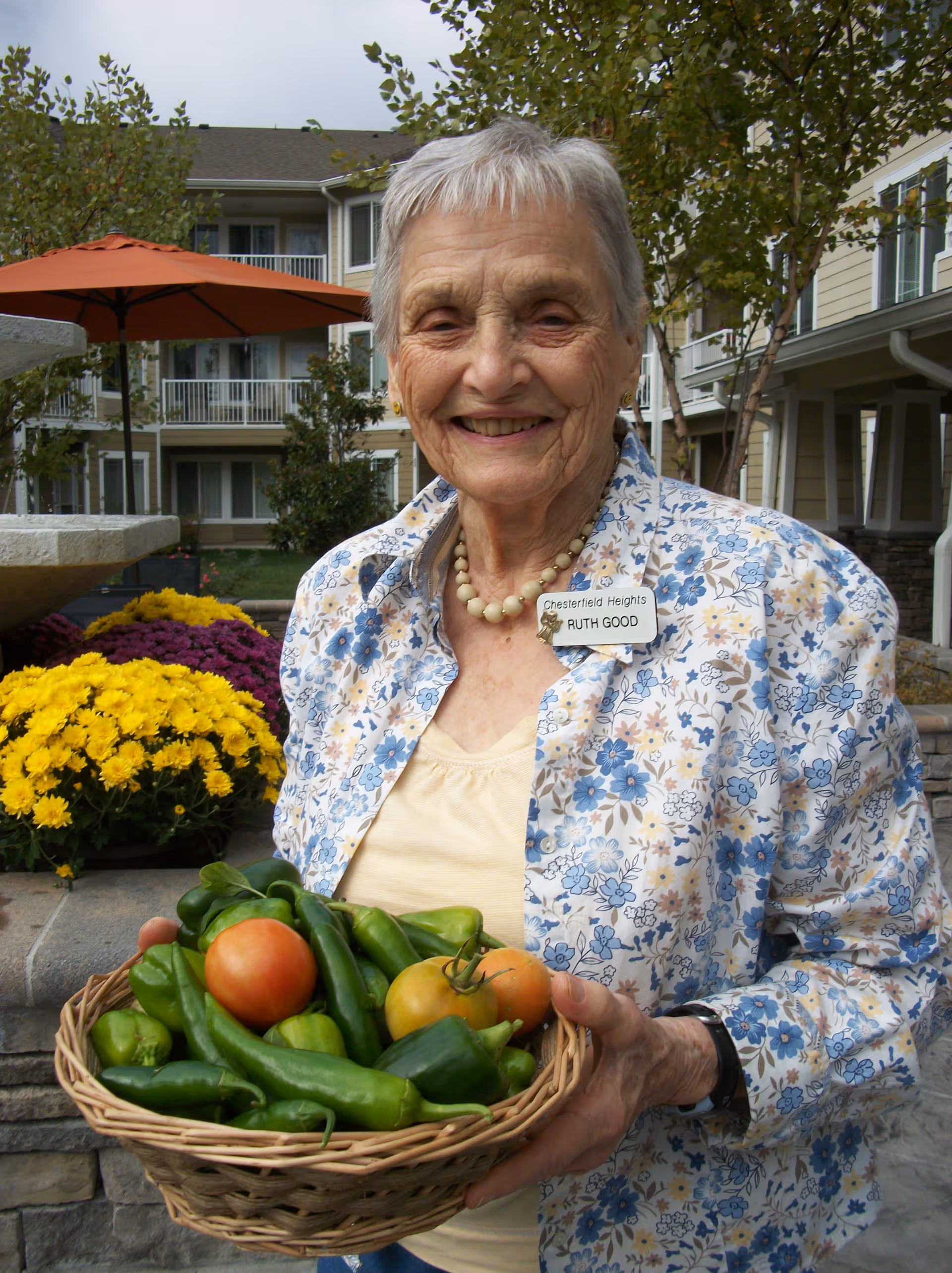 An elderly woman with short gray hair wearing a floral shirt and a name tag that reads 'Chesterfield Heights Ruth Good' is smiling and holding a basket filled with fresh green peppers and tomatoes. Behind her are yellow and purple flowers, a large orange umbrella, and a multi-story residential building with balconies.