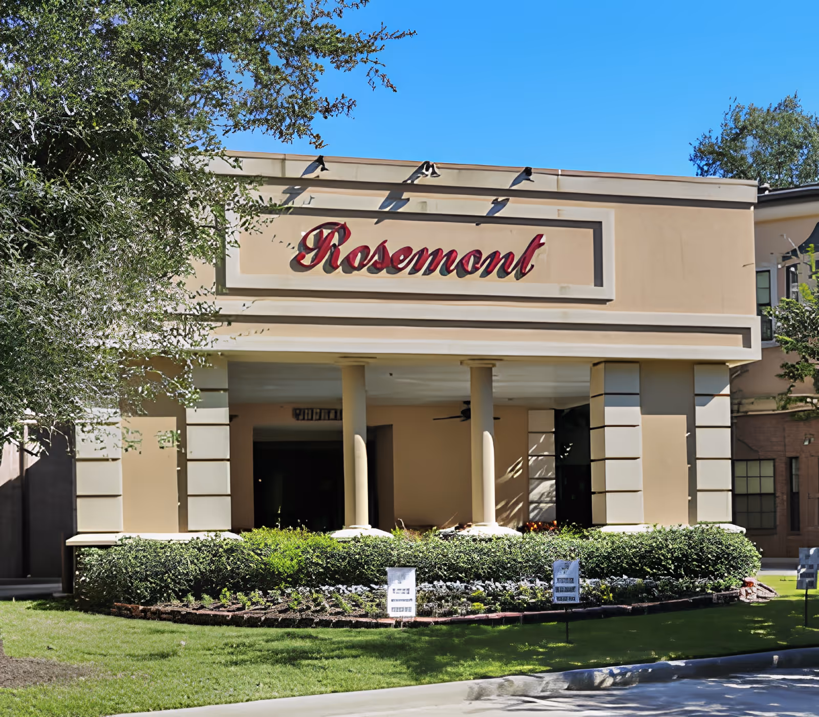 Exterior view of the Rosemont facility building with beige walls, columns, and a landscaped garden with green bushes and flowers in front under a clear blue sky.