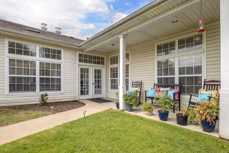Outdoor patio area at Brookdale Broken Arrow featuring a covered porch with white siding, multiple windows, a glass door, several black rocking chairs with colorful cushions, potted plants, and a green lawn in the foreground under a partly cloudy sky.