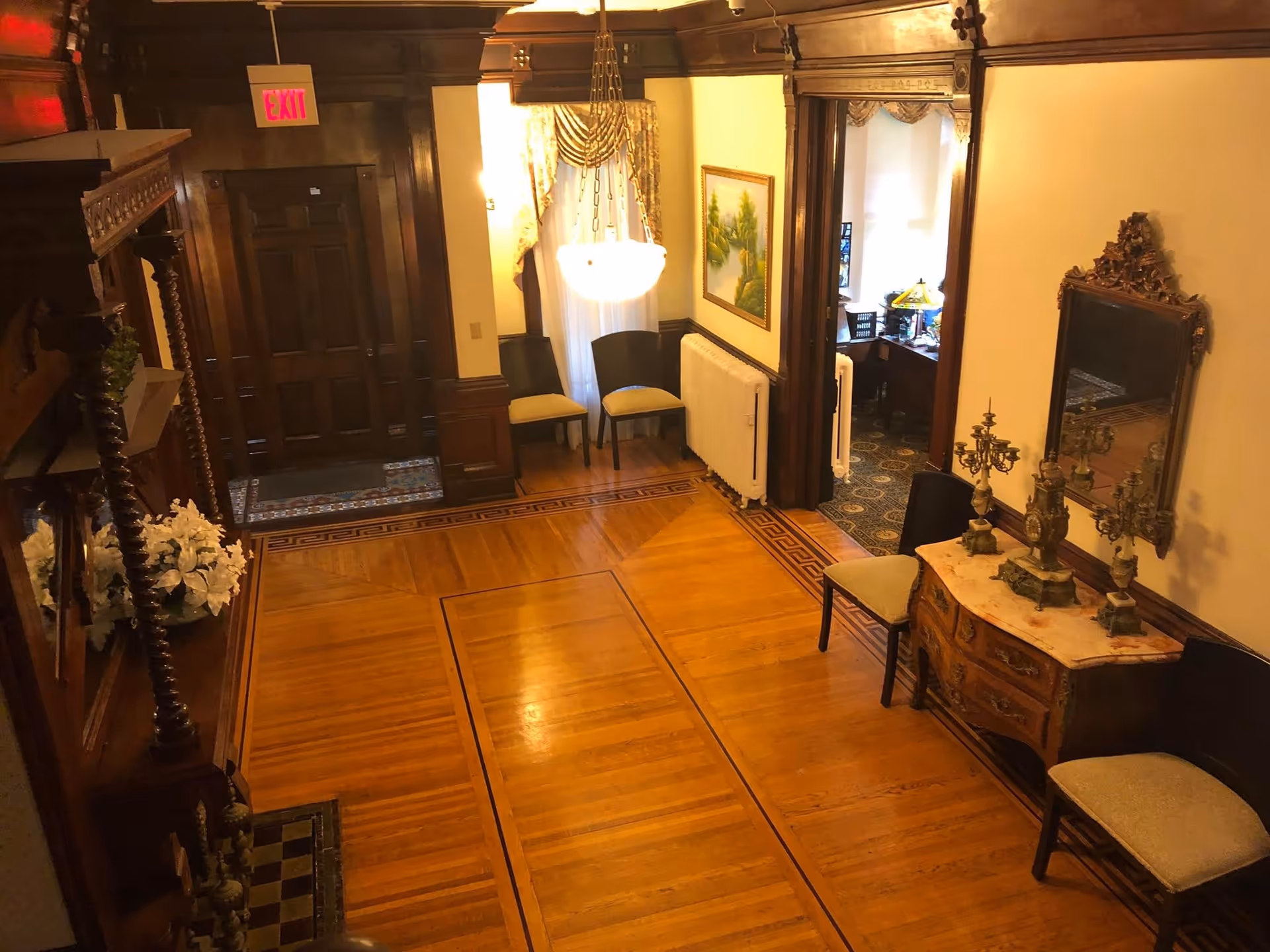 Antique-styled interior entry hall with polished hardwood floors, ornate wood trim, chairs, a console table with mirror and a hanging chandelier.