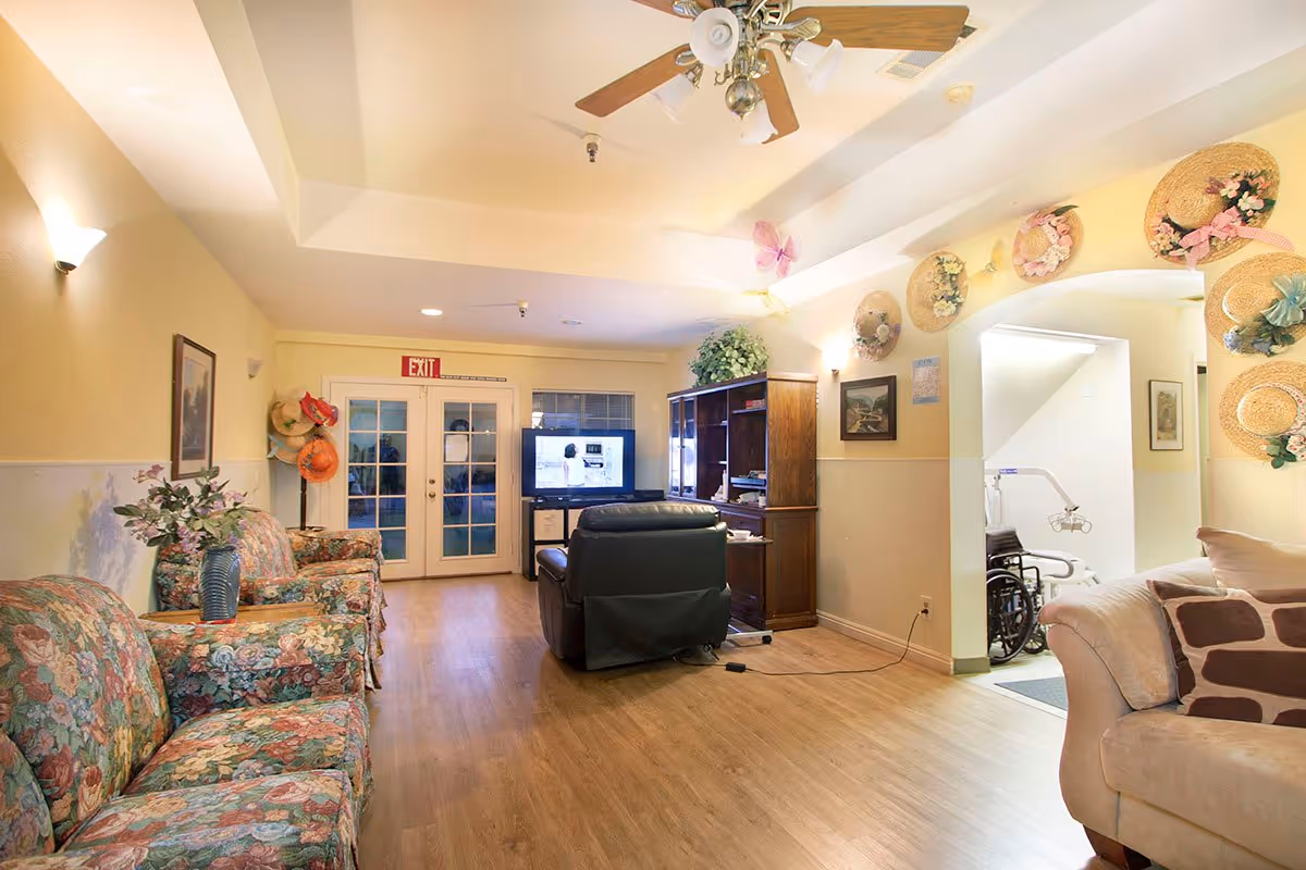 A cozy living room area in a senior living facility with floral patterned couches on the left, a beige couch with patterned pillows on the right, and a black recliner chair facing a TV on a wooden cabinet. The walls are decorated with straw hats adorned with flowers and butterflies. There is a ceiling fan with lights, wooden flooring, and a double glass door with an exit sign above it at the far end. A wheelchair and medical equipment are visible through an archway on the right.