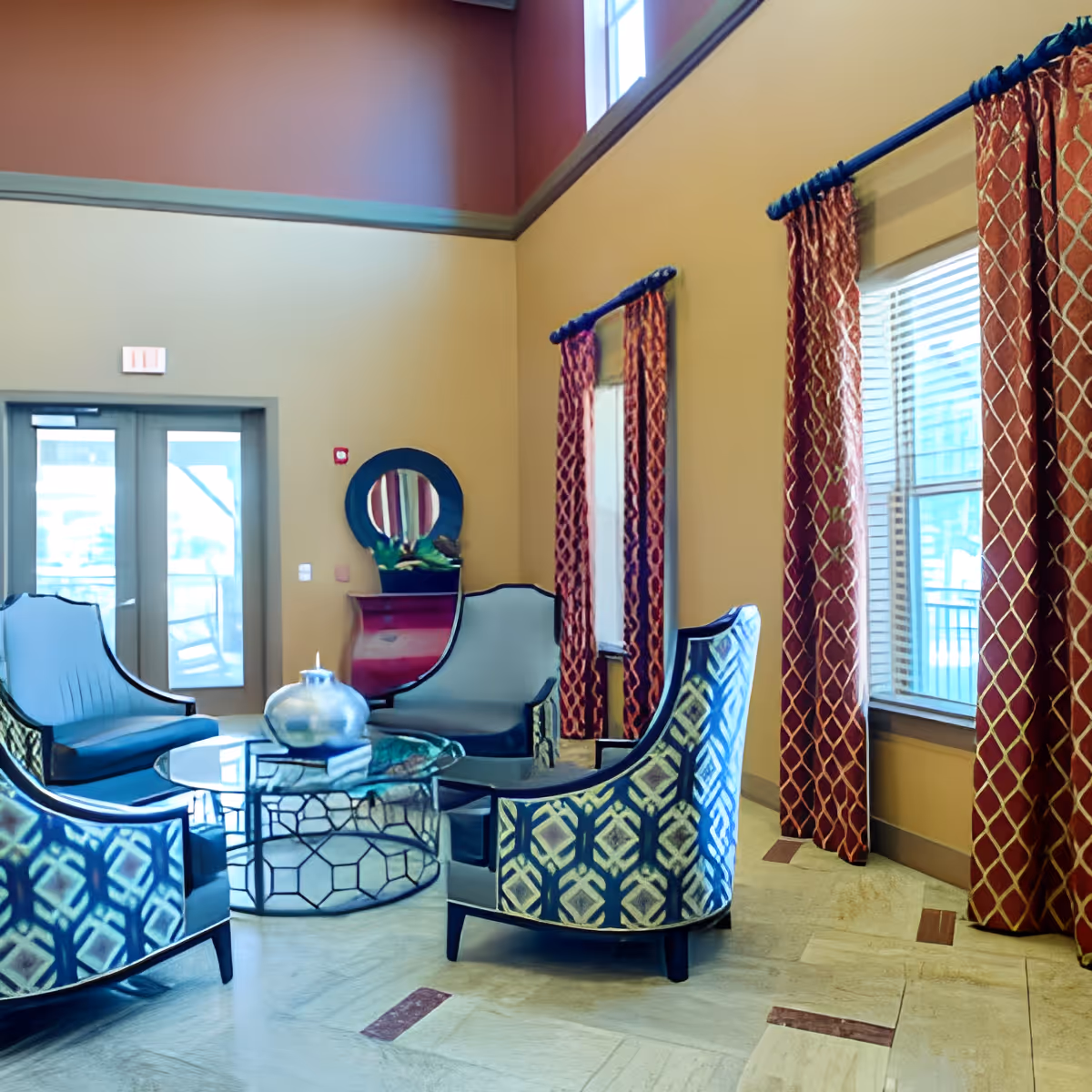 A cozy sitting area with four patterned armchairs arranged around a glass coffee table with a decorative vase. The room has tall ceilings, large windows with red patterned curtains, and a small red console table with a round mirror above it near a set of double glass doors.