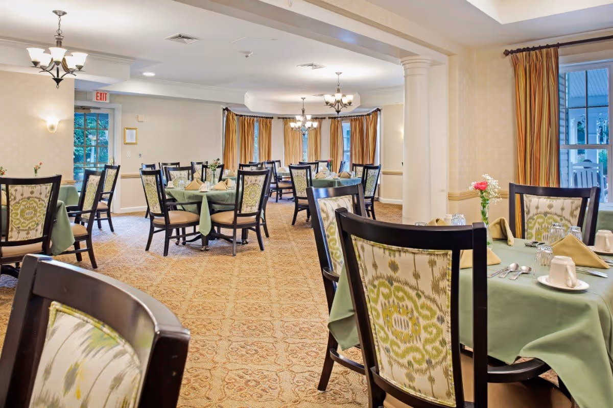 A dining room with multiple round tables covered with green tablecloths, each set with beige napkins, cups, glasses, and silverware. The chairs have dark wooden frames with patterned upholstery. The room has beige walls, carpeted floors, large windows with gold curtains, and ceiling light fixtures.