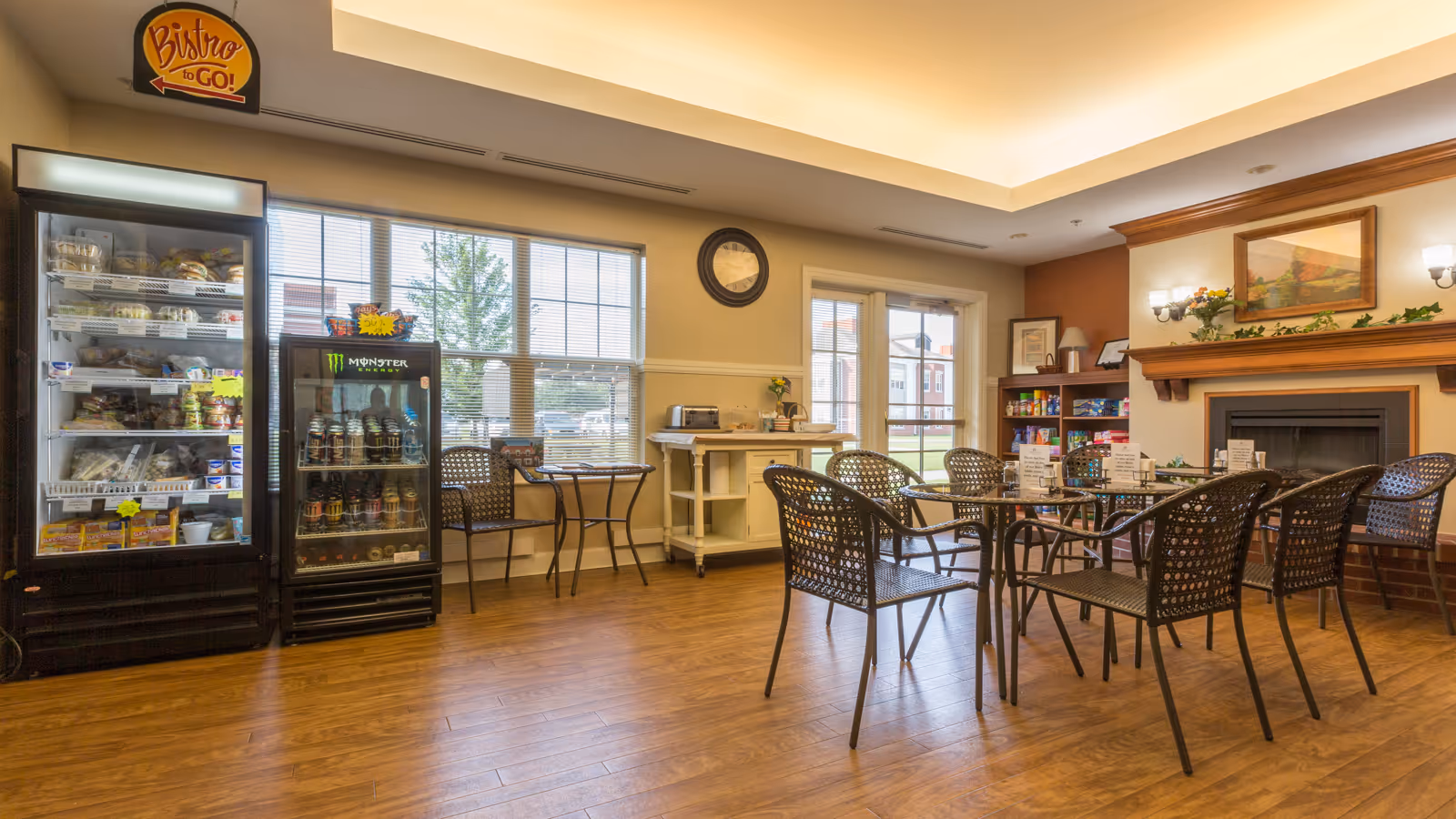 A cozy dining area in a retirement community with wooden flooring, several round tables with wicker chairs, a fireplace with a painting above it, and shelves with snacks and board games. There are two refrigerated display cases stocked with drinks and food items, and a sign above them reads 'Bistro to Go!'. Large windows and a glass door let in natural light.