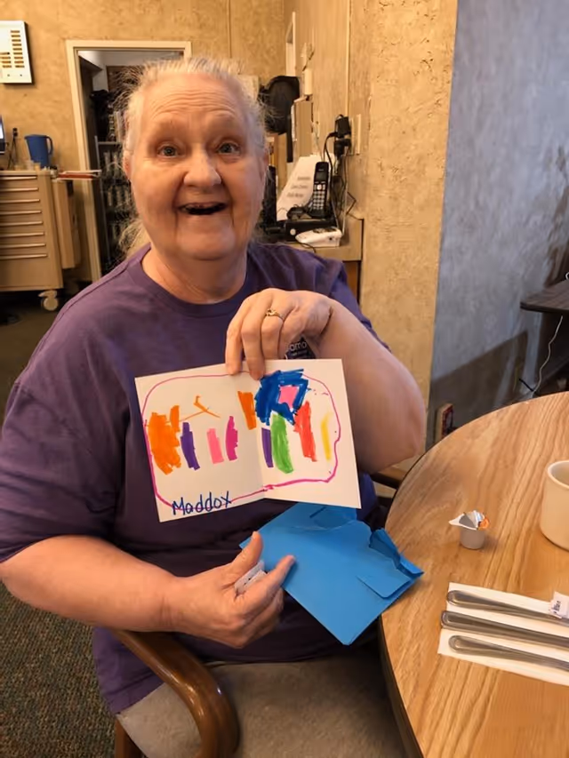 An elderly woman sitting at a round wooden table inside a room, smiling and holding up a colorful drawing with the name 'Maddox' written on it. She is wearing a purple shirt and appears happy. On the table are utensils wrapped in a napkin, a white cup, and a small container of creamer. The background shows a beige wall and some office equipment.