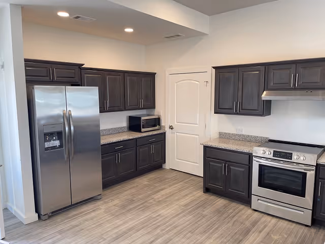 Modern kitchen with stainless steel refrigerator and oven, dark wood cabinets, granite countertops, and wood-look flooring.