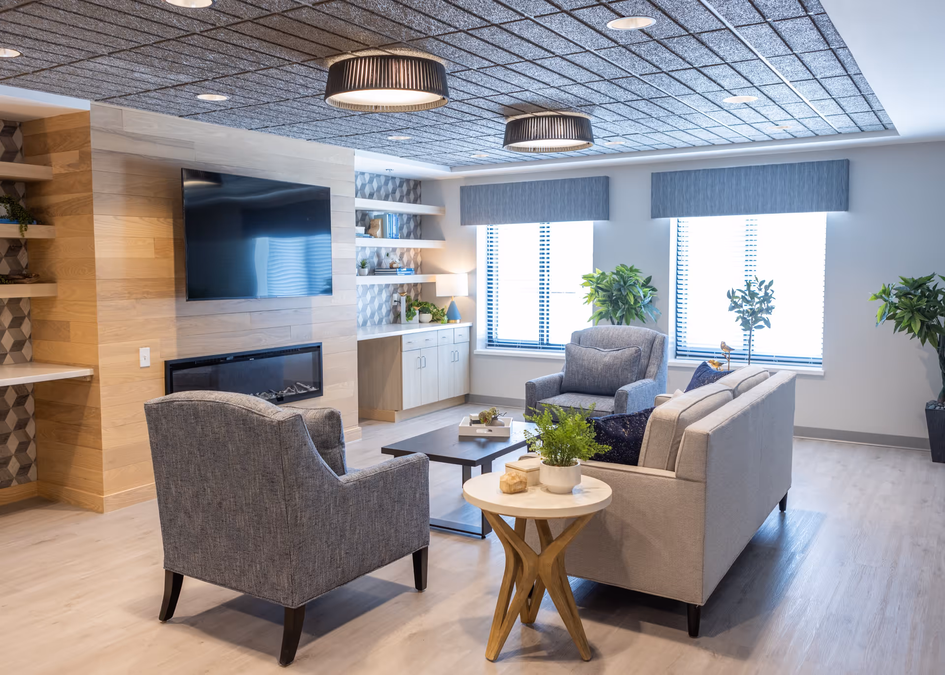A modern living room in a senior living facility with two gray armchairs and a light gray sofa arranged around a dark coffee table. There is a small round side table with a potted plant in the foreground. The room features large windows with blinds, built-in shelves with decorative items, a wall-mounted flat-screen TV above a fireplace, and ceiling lights with black shades.