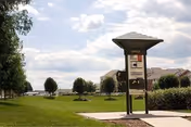 Grassy outdoor campus with a covered directory sign by a pathway, trees, and residential buildings under a partly cloudy sky.
