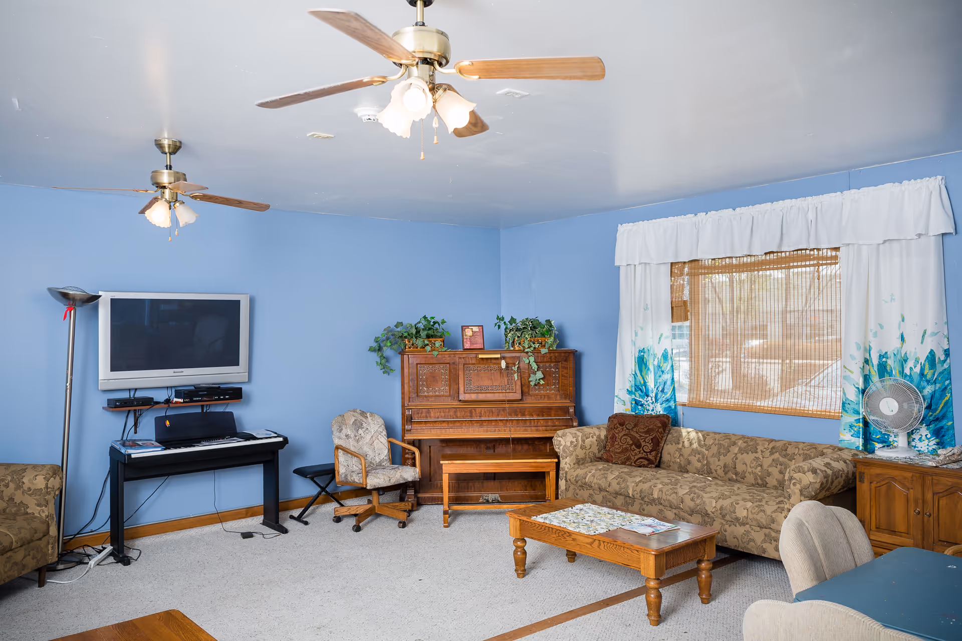 A cozy living room with light blue walls, two ceiling fans with lights, a wall-mounted flat-screen TV above a keyboard piano, a wooden piano with plants on top, a patterned armchair, a floral-patterned sofa, a wooden coffee table, a window with white and blue curtains, and a small fan on a wooden cabinet.