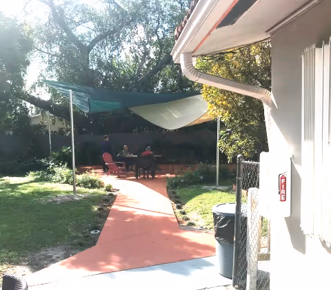 Outdoor seating area with two people sitting at a table under shade sails, surrounded by trees and greenery, with a red paved walkway leading to the seating area from a building corner.