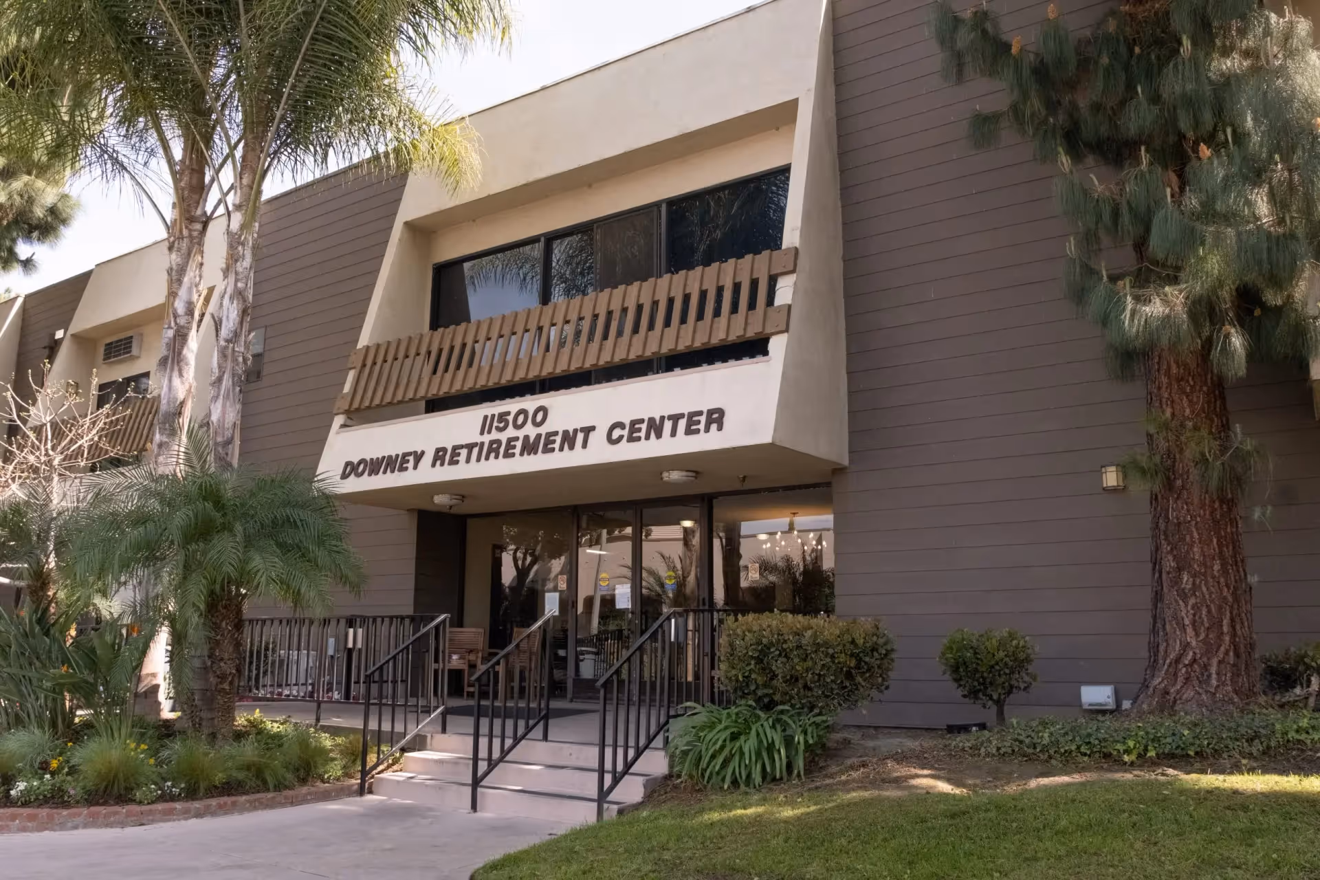 Exterior front view of Downey Retirement Center building with entrance steps, handrails, and surrounding greenery including palm trees and bushes.