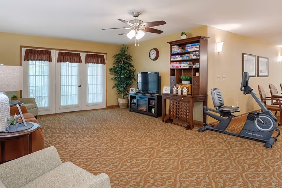 A spacious living room area with patterned carpet, a ceiling fan with lights, and large French doors with sheer curtains. The room features a TV on a stand, a tall wooden shelf filled with board games, a potted plant, and an exercise bike. There are also chairs and a table partially visible on the right side.