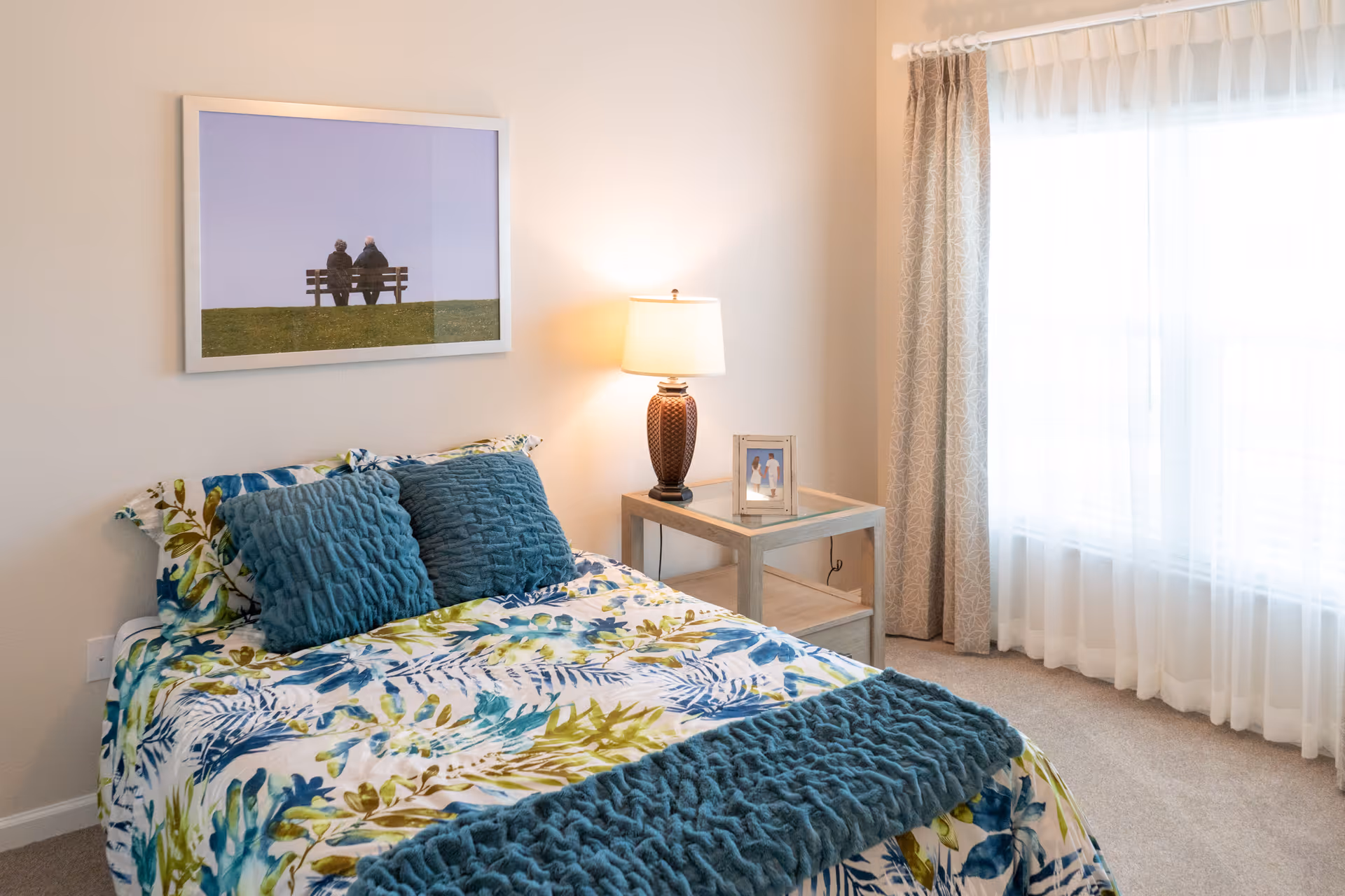 A bright bedroom with a bed covered in a floral patterned bedspread in shades of blue and green, two textured blue pillows, and a matching blue throw blanket. Next to the bed is a wooden nightstand with a lamp and a framed photo. A large window with sheer white curtains and patterned drapes lets in natural light. A framed picture of two people sitting on a bench hangs on the wall above the bed.