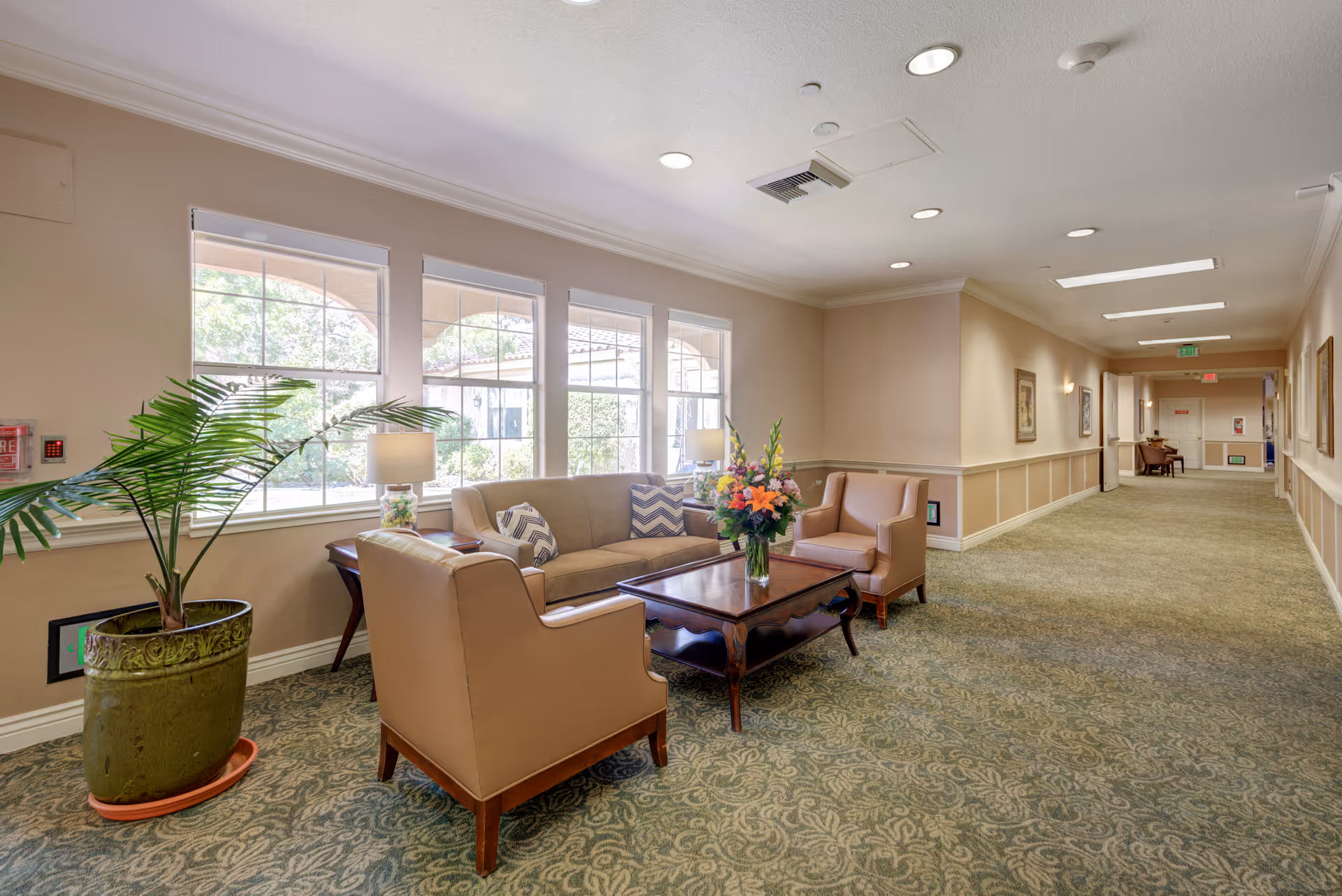 A bright and spacious hallway seating area in a senior living facility with large windows letting in natural light. The seating area includes a beige sofa, two beige armchairs, a wooden coffee table with a flower arrangement, a side table with a lamp, and a large potted plant. The hallway is carpeted with a patterned design and has framed artwork on the walls.