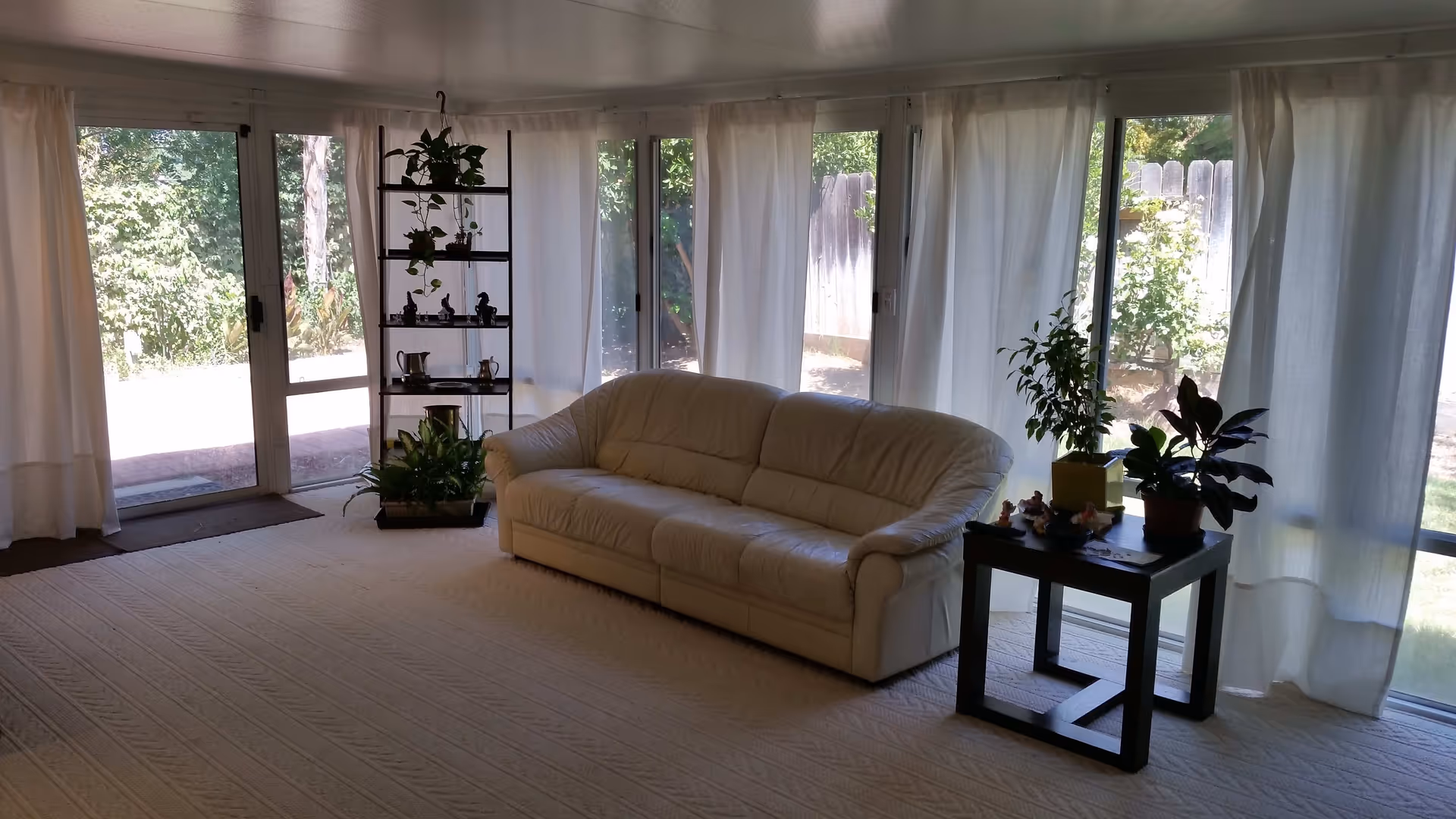 Bright living room with a cream leather sofa, side table and plants in front of floor-to-ceiling windows with sheer curtains.