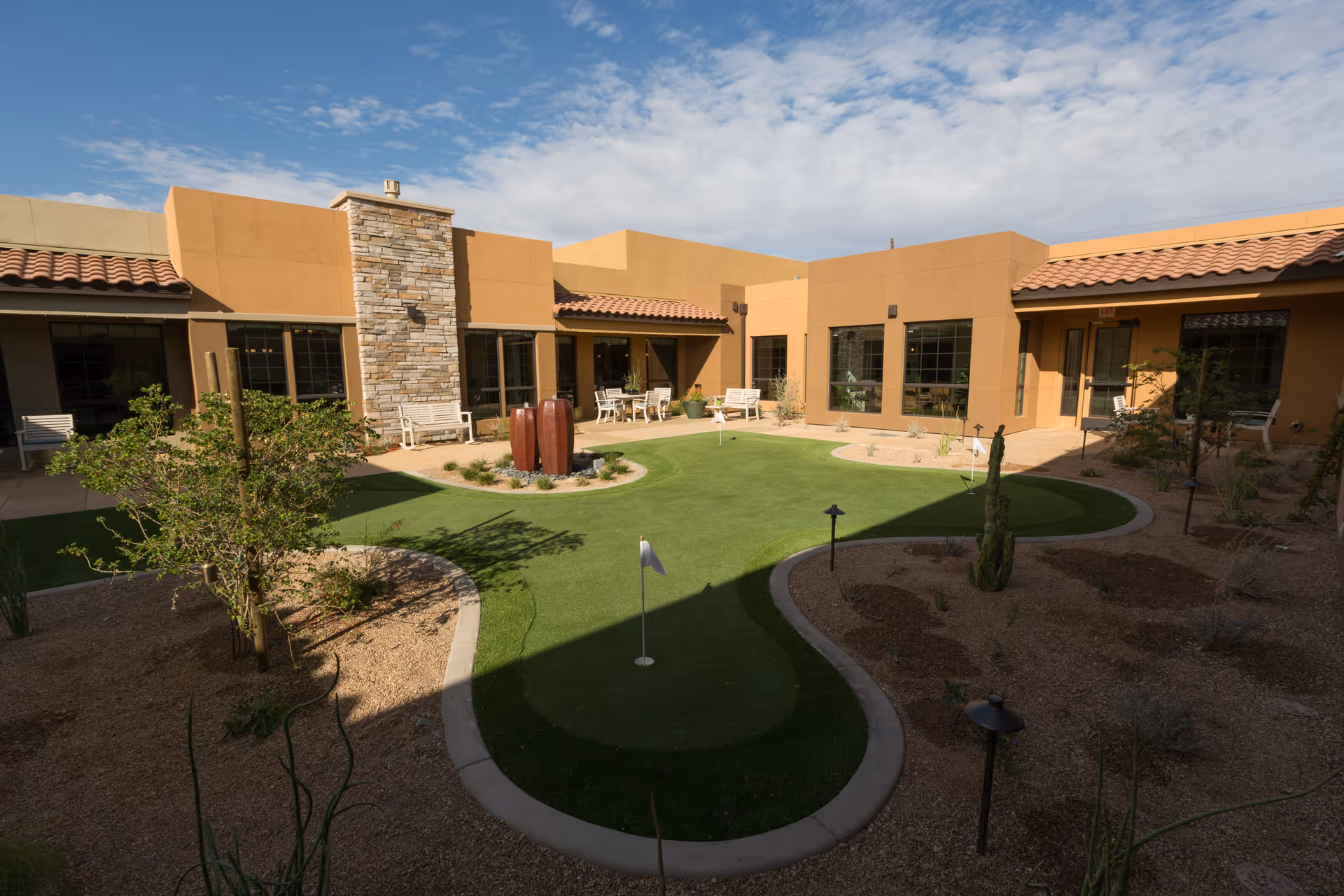 Outdoor courtyard area at Lone Mountain Memory Care featuring a putting green with small flags, surrounded by desert landscaping with gravel and sparse plants. The courtyard is enclosed by a single-story building with tan stucco walls, large windows, and a stone chimney. Several benches and chairs are placed along the building walls under covered walkways.