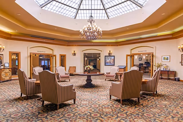 A spacious, elegant common area with a large round table in the center surrounded by eight striped upholstered armchairs. The room features a patterned carpet, a chandelier hanging from a large skylight ceiling, and doorways labeled 'Heritage Room' and 'Bennett Cafe'. The walls are decorated with framed artwork and sconces.