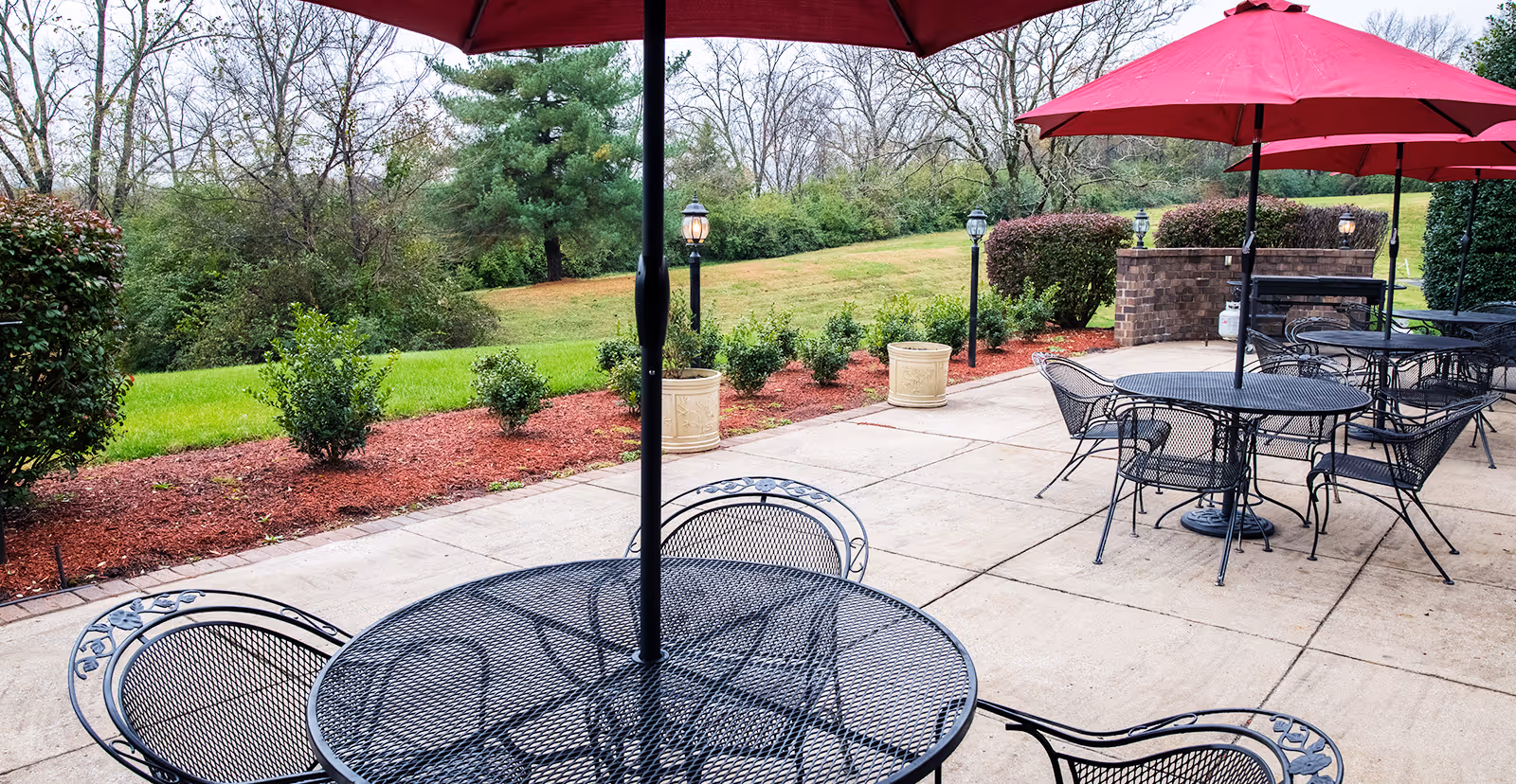 Outdoor patio with metal tables and red umbrellas overlooking a grassy lawn and landscaped shrubs.