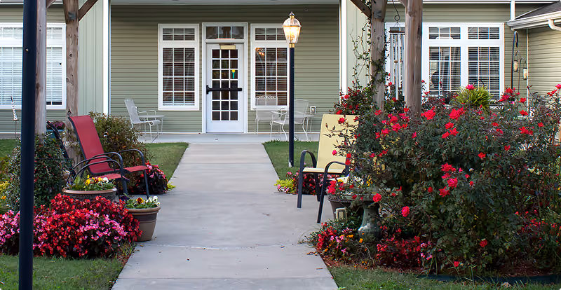 Outdoor garden area with a concrete walkway leading to a building entrance with a glass door and windows. The garden features colorful flowers, potted plants, and several chairs including red and yellow cushioned ones. There are wooden pergolas and a lamp post along the walkway.