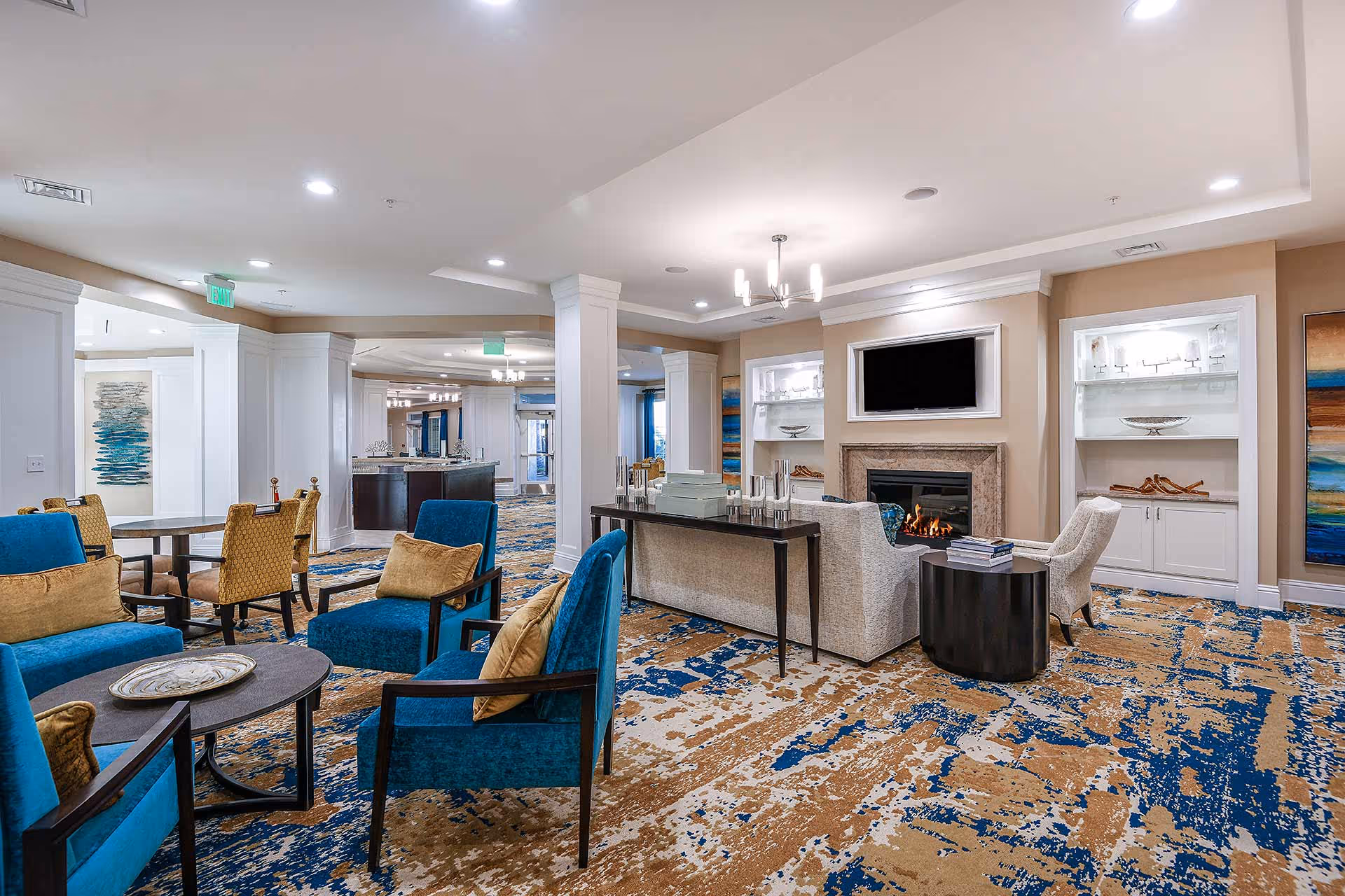 A spacious, well-lit living room area in a senior living facility with blue and beige patterned carpet, blue armchairs with tan cushions, beige armchairs around a fireplace with a mounted TV above it, and tables with decorative items. The room has white columns, built-in shelves with glassware, and modern ceiling lights.