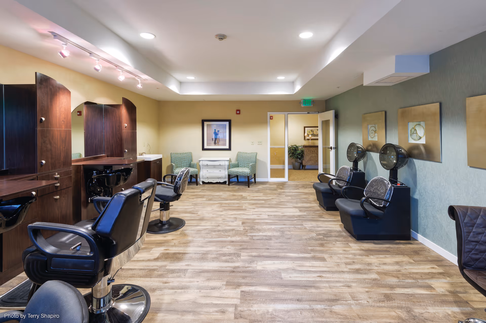 Interior view of a senior living facility's hair salon with multiple black salon chairs and hair washing stations on the left, two hair drying chairs on the right, wooden flooring, green and beige walls, and two green patterned chairs with a small white dresser at the back near a framed picture.