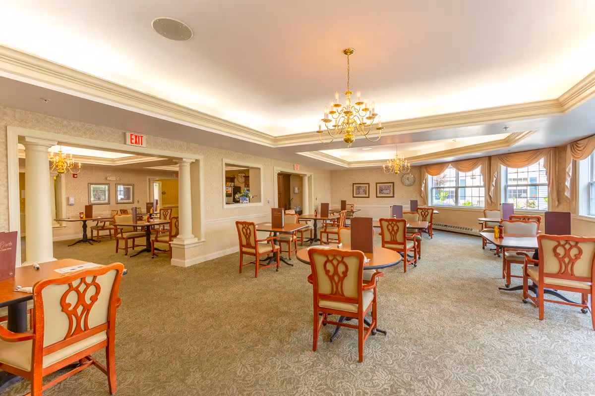 A spacious dining room in a senior living facility with multiple round tables and wooden chairs with cushioned seats. The room features large windows with draped curtains allowing natural light to fill the space. Elegant chandeliers hang from the ceiling, and there are decorative columns and framed artwork on the walls. Menus or informational cards are placed on each table.