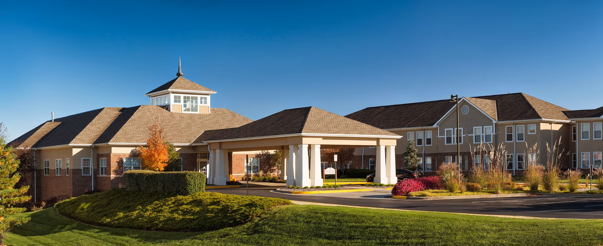 Front exterior of a senior living facility with a covered porte-cochère, landscaped lawns, and a clear blue sky.