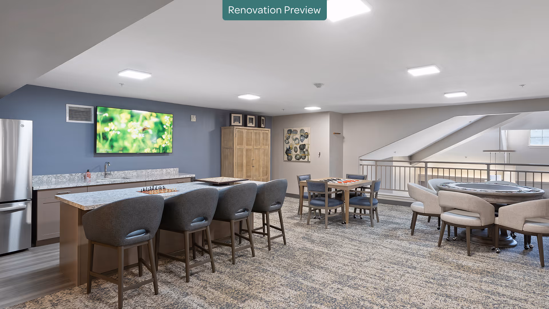 A spacious, modern common area with a kitchenette featuring a refrigerator, sink, and marble countertops. There are four gray cushioned bar stools at the kitchen island, a wall-mounted TV displaying a nature scene, and several tables with chairs arranged for games or socializing. The room has carpeted flooring, neutral-colored walls, and bright ceiling lights. A sign at the top center reads 'Renovation Preview.'