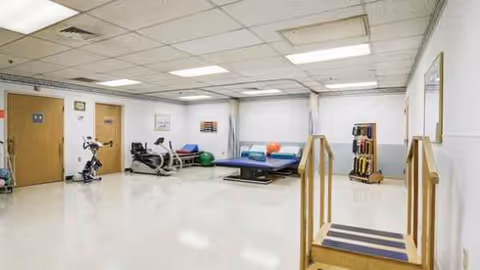Indoor rehabilitation gym with exercise bikes, weights, a therapy bed, and a small stair ramp.