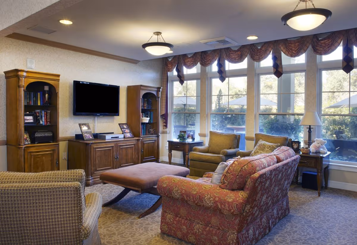A cozy living room with large windows letting in natural light. The room features a patterned red sofa, two armchairs, a large ottoman, wooden cabinets with books and decorative items, a flat-screen TV mounted on the wall, and a side table with a lamp and a teddy bear. The windows have valance curtains and outside greenery is visible.