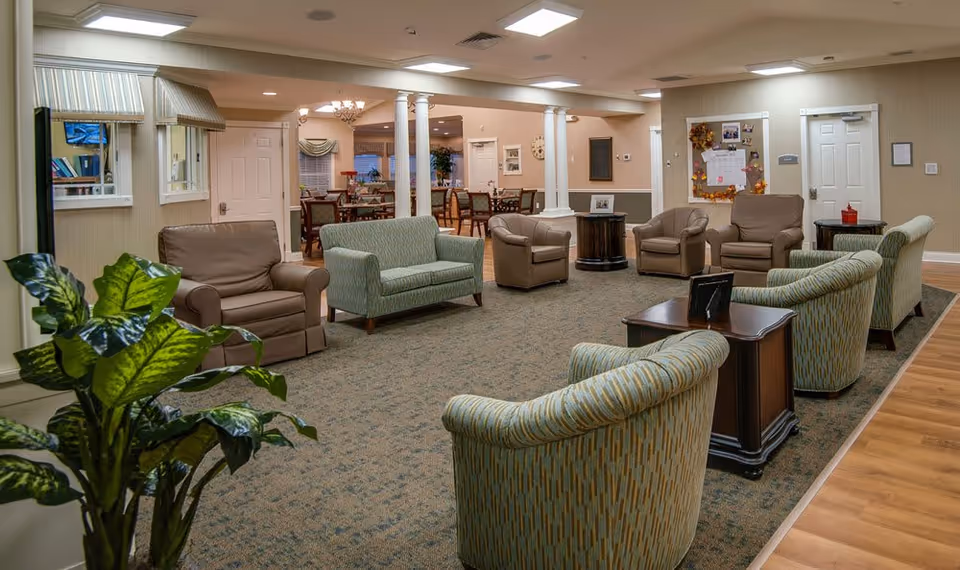 Spacious senior living common lounge with multiple upholstered chairs and sofas arranged around small tables and a dining area visible in the background.