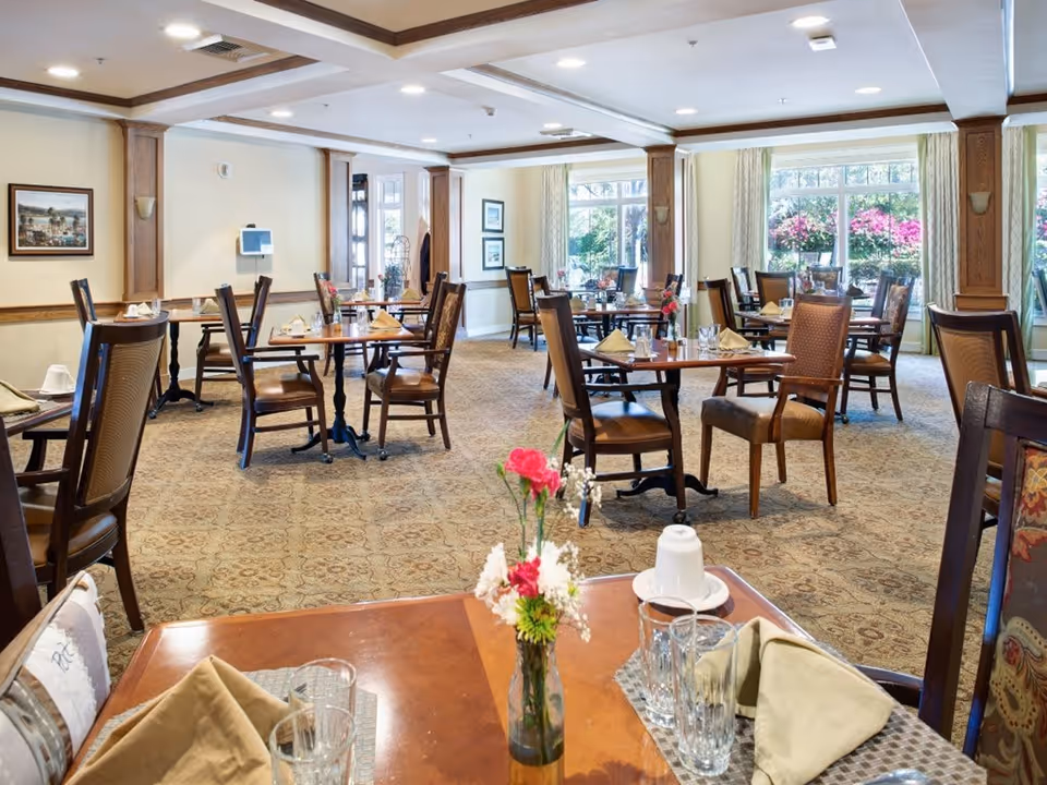 A bright and spacious dining room with multiple wooden tables and chairs arranged neatly. Each table is set with napkins, glasses, and small flower vases. Large windows on one side let in natural light and offer a view of colorful flowers outside. The room has a patterned carpet and light-colored walls with wooden trim and framed artwork.