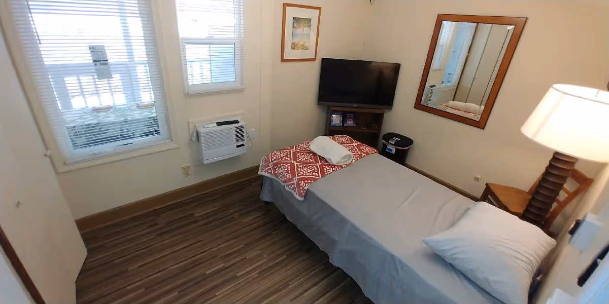 A small bedroom with a single bed covered with a gray sheet and a red patterned blanket at the foot. A white towel is folded on the blanket. The room has wooden flooring, a wall-mounted air conditioner below two windows with blinds, a flat-screen TV on a small stand, a wooden chair, a lamp with a white shade, a framed picture on the wall, and a large mirror.