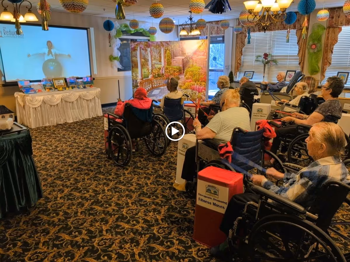 A group of elderly individuals in wheelchairs participating in a seated exercise session in a decorated room. They are using drumsticks to play on red and white boxes labeled 'Therapeutic Drumming Fitness Moves.' A large screen at the front of the room shows an instructor demonstrating exercises with a fitness ball. The room has patterned carpet, hanging decorations, and windows with blinds.