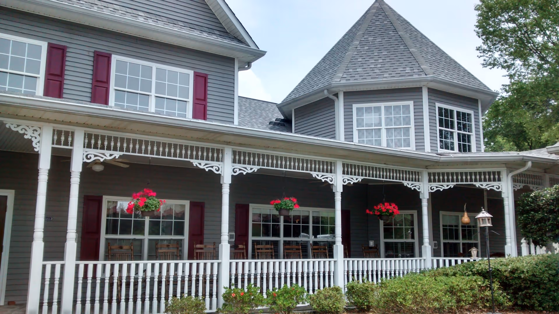 Exterior view of a senior living facility with gray siding, white trim, and red shutters. The building features a wrap-around porch with white railings and decorative trim, hanging flower baskets with red flowers, rocking chairs, and greenery in front.