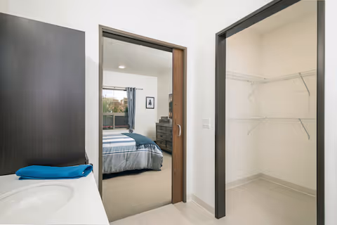 Interior view of a senior living suite showing a bathroom vanity and sink, an open walk-in closet, and a doorway leading to a bedroom with a bed and dresser.