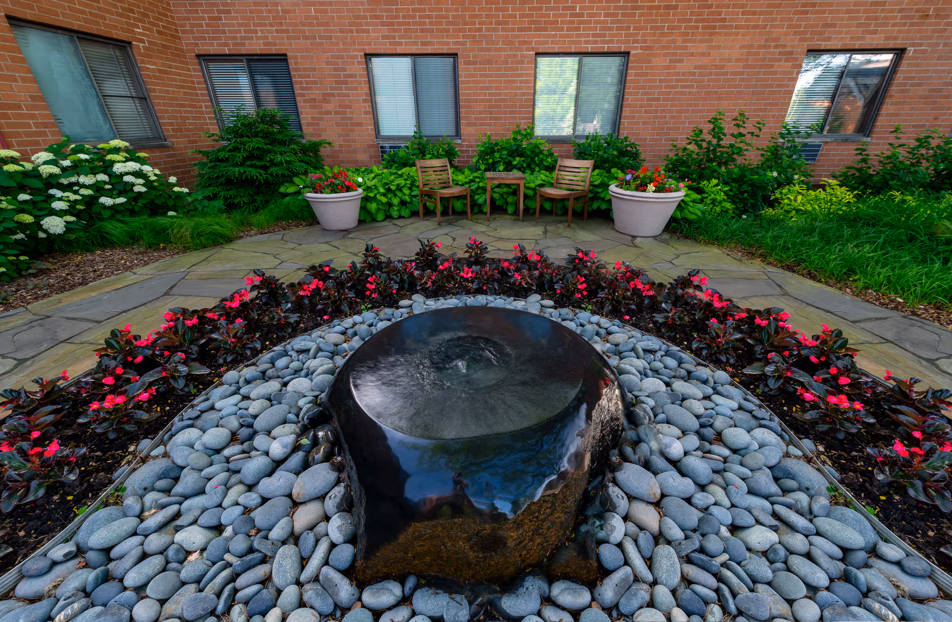 A peaceful outdoor courtyard area with a circular stone water feature surrounded by smooth gray pebbles and red flowering plants. In the background, there are two wooden chairs and a small table placed on a stone-paved patio, with green shrubs and plants along the brick building wall with several windows.