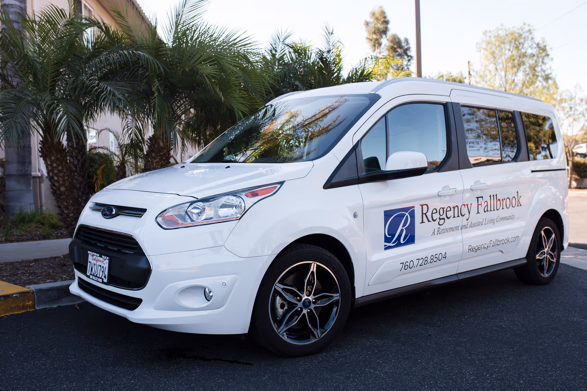 White Ford van parked on a street with palm trees in the background. The van has signage on the side that reads 'Regency Fallbrook, A Retirement and Assisted Living Community' along with a phone number and website.