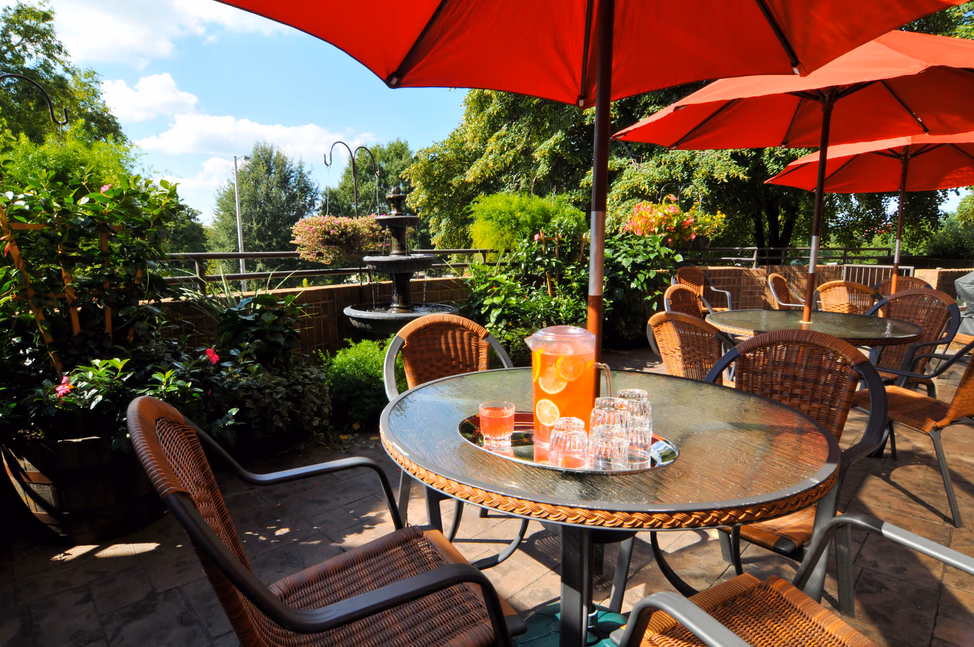 Outdoor patio area with round glass tables and wicker chairs under large red umbrellas. One table has a pitcher of iced tea with lemon slices and several empty glasses on a tray. The patio is surrounded by lush green plants and trees, with a multi-tiered water fountain in the background under a blue sky with some clouds.