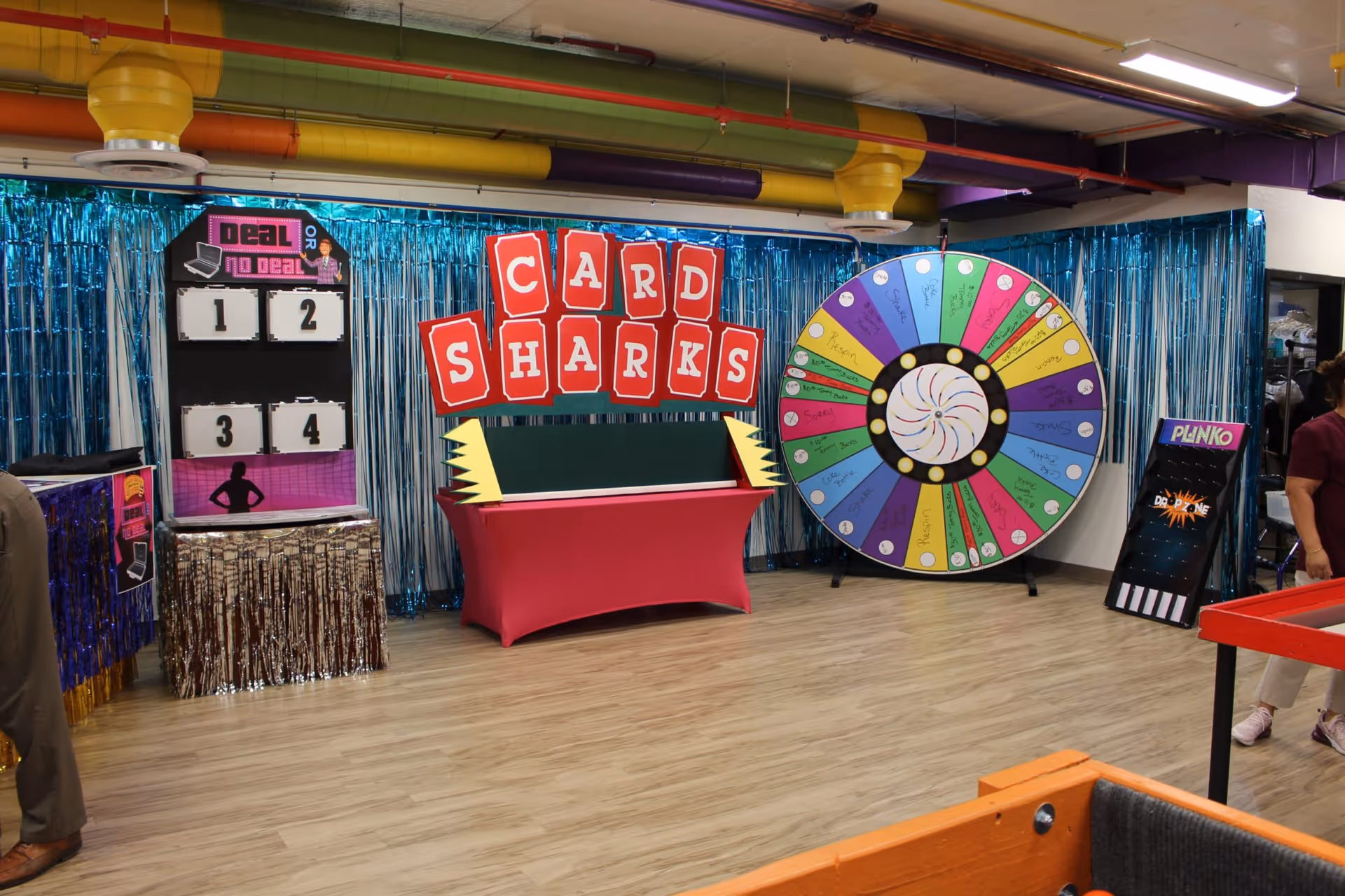 A colorful indoor game area with a Deal or No Deal game board, a large spinning prize wheel, and a Card Sharks sign above a table covered in a red cloth. The background features shiny blue streamers and exposed colorful pipes on the ceiling. A person is partially visible on the right side of the image.